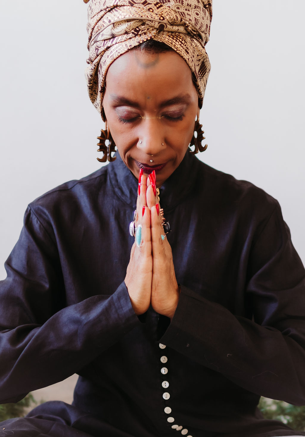 Person in patterned headwrap and dark tunic sits with eyes closed, palms pressed together in prayer at heart-center.