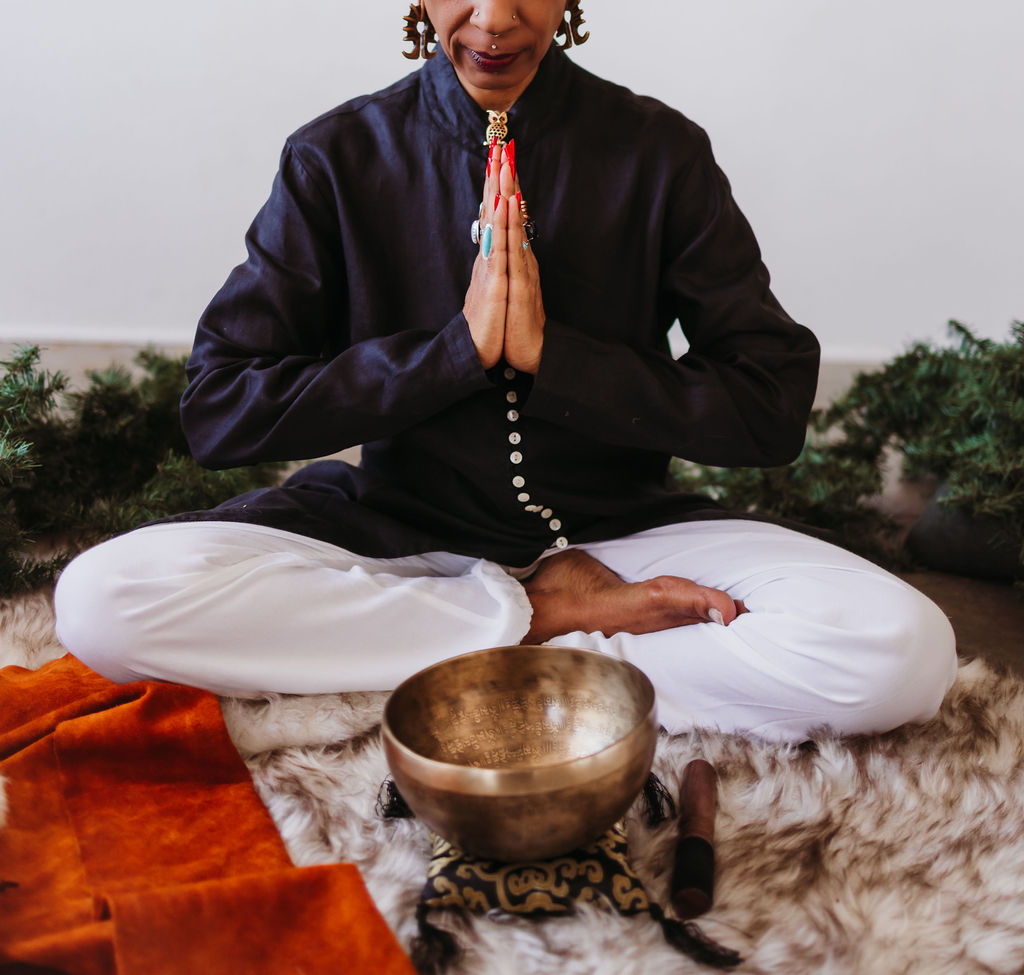 Person sitting cross-legged on a soft fur rug, hands pressed together at heart-center, with a bronze singing bowl and mallet in front and evergreen branches nearby.